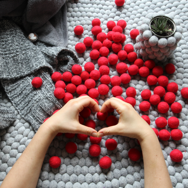Czerwone kulki filcowe na tle szarego dywanu. Ręce ułożone w znak serca