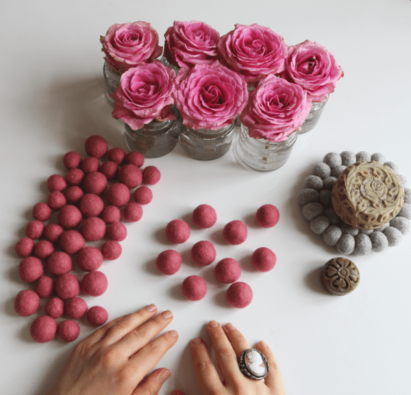 Kulki filcowe w kolorze różowym na białym tle obok kwiatów róży. Widoczne ręce i pierścionek na palcu.